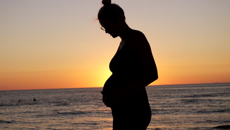 Happy-smiling-pregnant-woman-watching-belly-and-touching-in-front-of-peaceful-ocean-at-sunset---Silhouette-of-female-person-at-beach---close-up-slow-motion