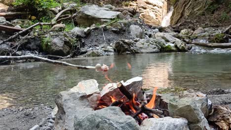 Guy-cook-sausages-on-a-stick-on-camp-fire-with-waterfall-and-river-in-background