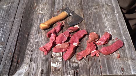 Sharp-small-hiking-axe-thrown-down-at-table-besides-sliced-raw-deer-meat-in-slow-motion---Outdoor-food-preparation-and-survival