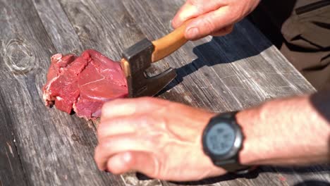 Male-hands-using-small-hiking-axe-to-cut-filets-from-raw-meat-outdoor-on-wooden-table---Food-preparation-in-nature