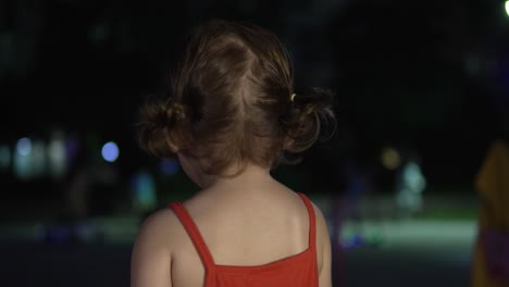 View-Behind-Beautiful-Toddler-Holding-Food-And-Watching-People-In-The-Park-At-Nighttime