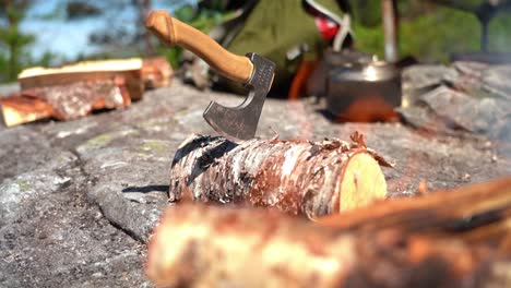 Hiking-axe-inserte-in-a-birch-log-with-bonfire-and-flames-in-blurred-out-foreground---Handheld-slow-motion-hiking-clip-from-forest