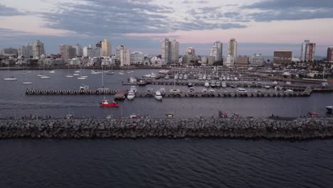 Cinematic-aerial-tracking-shot-of-red-fishing-motorboat-entering-the-city-harbor-of-Punta-del-Este-in-Uruguay-with-skyline-and-skyscrapers-in-background