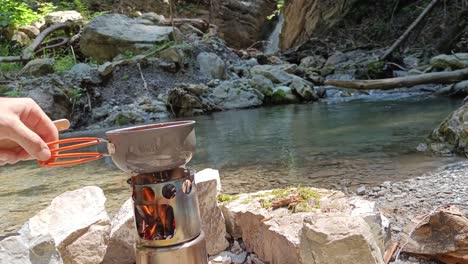 Guy-put-pot-with-soup-on-hobo-cooker-and-stir-it-with-waterfall-and-river-in-background
