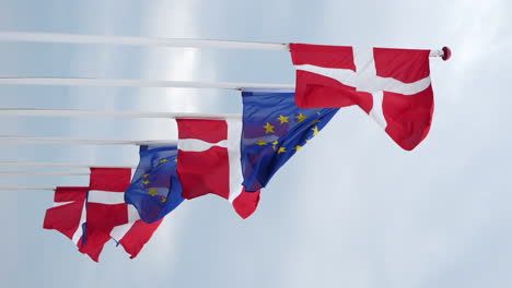 Vertical-Shot-Of-Danish-And-European-Union-Flags-On-Flagpoles-Flying-With-The-Wind