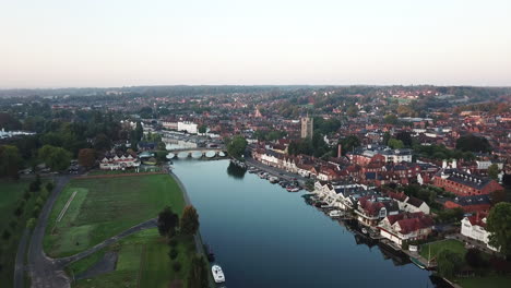 Beautiful-morning-drone-shot-of-Henley-on-Thames,-Oxfordshire,-panning-along-the-regatta-stretch-of-the-river-and-flying-towards-the-bridge