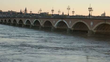Brücke-Mit-Einem-Reißenden-Fluss-Un-Ein-Sonnenuntergang-Mit-Orangefarbenem-Hintergrund-Und-Straßenlaternensilhouetten-In-Budeaux,-Frankreich