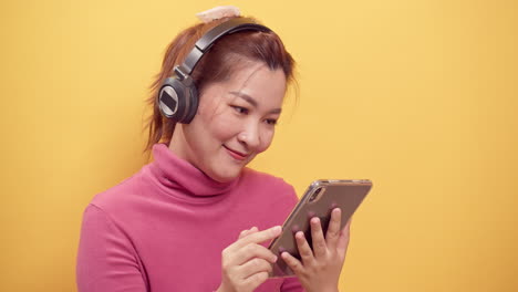 Wunderschöne-Asiatische-Junge-Frau-Der-Schönheit,-Die-Ein-Digitales-Tablet-Und-Eine-Streaming-anwendung-Für-Das-Fröhliche-Hören-Von-Musik-über-Kopfhörer-Mit-Entspanntem-Auf-Hellgelbem-Hintergrund-Verwendet-1