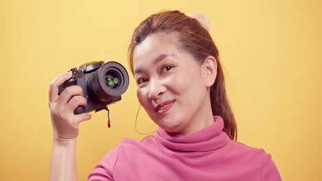 Junge-Asiatische-Frau,-Die-In-Rosa-Kleidung-Mit-Einer-Digitalkamera-Vor-Einem-Isolierten-Gelben-Hintergrund-Mit-Kopierraum-Für-Werbung-Spielt