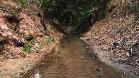 Deep-sandy-ditch-with-flowing-water-in-dense-Vietnamese-jungle,-fly-forward-view