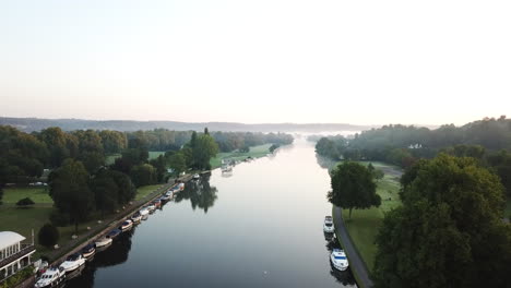 Beautiful-drone-shot-of-Henley-on-Thames-at-sunrise,-flying-along-the-river-to-the-East-with-a-light-mist-over-the-river-and-surrounding-hills-and-fields
