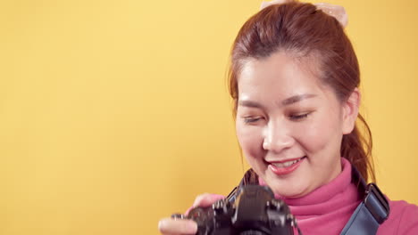Young-Asian-woman-playing-in-pink-clothing-using-a-digital-camera-against-an-isolated-yellow-background