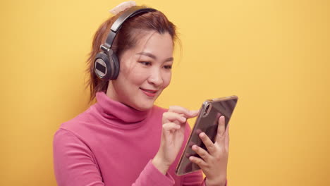 Lächelnde-Asiatische-Frau,-Die-Ein-Digitales-Tablet-Und-Eine-Streaminganwendung-Verwendet,-Die-Online-Musikentspannungen-Mit-Kopfhörern-Auf-Hellgelbem-Hintergrund-Hört
