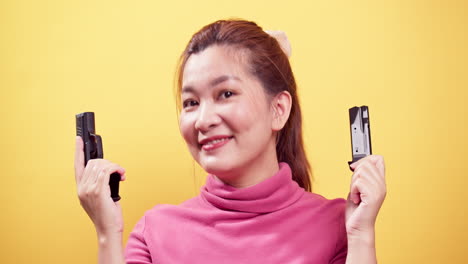 Nahaufnahme-Lächelnde-Asiatische-Frau,-Die-Pistole-Zur-Sicherheit-Auf-Munition-Auf-Hellgelbem-Hintergrund-überprüft