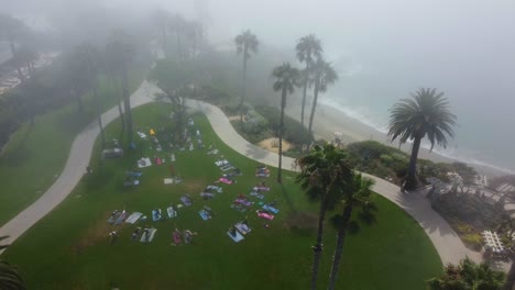 Birds-Eye-Aerial-View-of-People-at-Beachfront-Park-Grass-Having-Morning-Exercise-Under-Mist,-Drone-Shot