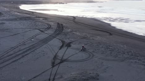 Motorcycle-rider-speeding-away-spraying-black-sand-on-beach-at-sunrise,-remote-volcanic-beach,-extreme-sports-in-nature