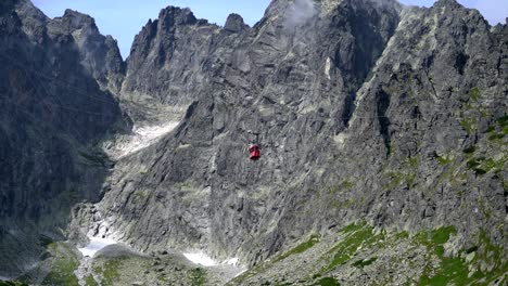 A-tiny-red-gondola-slowly-descends-in-front-of-rugged-jagged-cliffs,-The-High-Tatra-Mountains