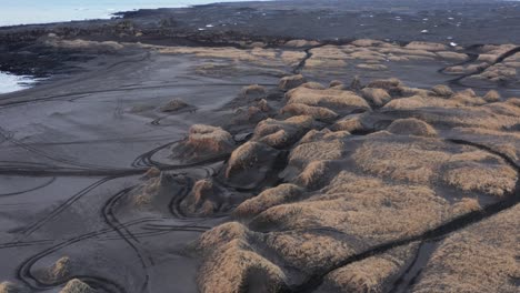 Sandvik-black-volcanic-sand-beach-with-motorcross-tire-tracks-and-grassy-dunes,-aerial