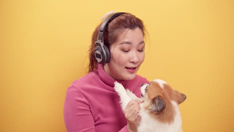 Wunderschöne-Asiatische-Frau-Verwendet-Ein-Digitales-Tablet-Und-Eine-Streaming-anwendung,-Um-Fröhlich-Musik-über-Kopfhörer-Zu-Hören,-Während-Sie-Den-Hund-Zur-Entspannung-Auf-Hellgelbem-Hintergrund-Spielt