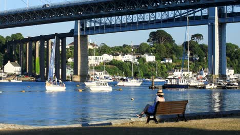 A-Lady-with-a-Hat-Sits-on-the-Bench-Overlooking-the-River-Tamar-with-the-Tamar-and-Royal-Albert-Bridge-in-the-Background-on-a-Summers-Evening-in-Saltash,-Cornwall