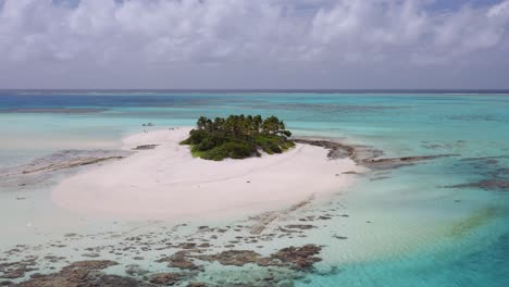 Anti-clockwise-orbit-of-small-sandy-island-with-palm-trees-surrounded-by-clear,-shallow-blue-water-and-coral-reef