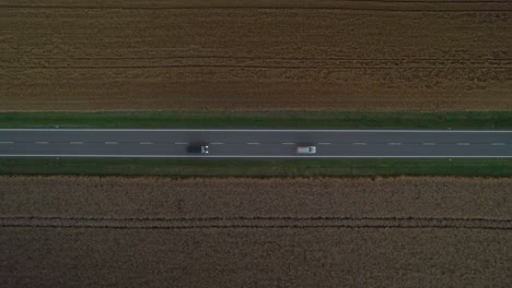 Cars-passing-by-with-their-headlights-on-on-a-straight-road-between-fields