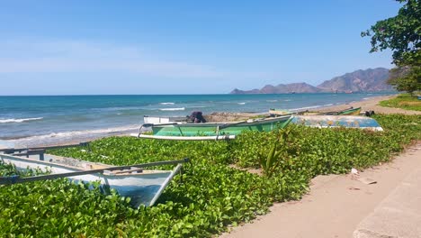 Colourful-Asian-traditional-wooden-fishing-canoes-lined-up-along-shoreline-with-ocean-waves-rolling-in-on-sunny,-blue-sky-day-in-capital-Dili,-Timor-Leste,-Southeast-Asia