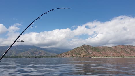 A-fishing-rod-with-line-out-trolling-for-fish-in-vast-calm-ocean-on-perfect-sunny,-blue-sky-summers-day-with-land-visible-in-the-distance