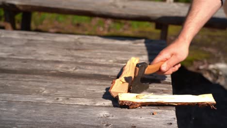 Male-hand-splitting-wood-with-small-hiking-axe-slow-motion---Sunny-day-outdoor-clip-with-firewood-standing-on-wooden-table-before-beeing-split-into-two-pieces
