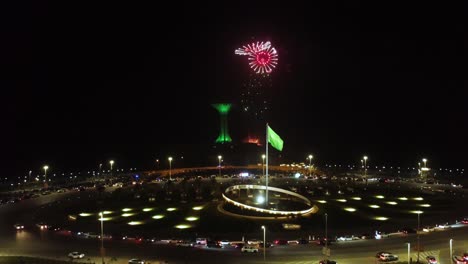 Aerial-circling-Saudi-Arabian-flag-fluttering,-fireworks-night,-Khobar-Tower-in-background