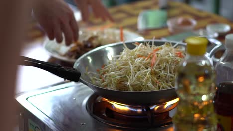 Person-Putting-Handfuls-of-Vegetables-into-a-Wok-on-a-Stove-High-Heat-to-Cook-Pad-Thai-in-an-Outdoor-Kitchen-in-Bangkok-Thailand-SLOW-MOTION