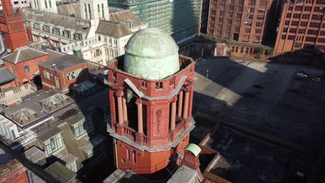 Aerial-drone-flight-over-Oxford-Road-showing-the-Refuge-Buildings-smaller-green-dome-alongside-the-rooftops-and-architecture-below-in-Manchester-City-Centre