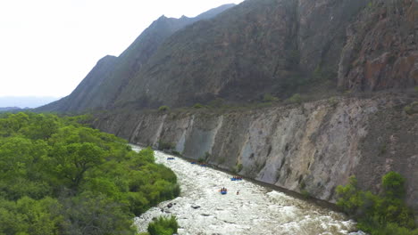 AERIAL---Epic-shot-of-kayaks-on-a-river-next-to-high-mountain-cliffs,-forward