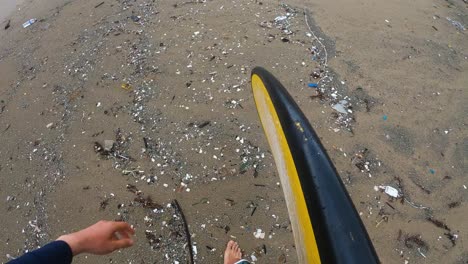Surfer-walks-along-sandy-beach-covered-in-micro-plastic-garbage-and-other-trash