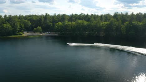 Aerial-view-of-a-woman-wakeboarding-or-water-skiing-speedily-on-a-lake-at-mid-day-with-many-beautiful,-green-trees-in-the-background