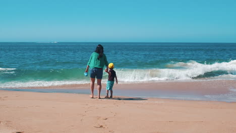 Back-view-of-a-latino-mother-with-her-son-in-the-beach-looking-at-the-sea-waves-during-summer-vacation