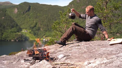 Man-sitting-down-on-rock-an-pouring-himself-a-cup-of-freshly-brewed-coffee-with-mountain-scenery-in-background-and-bonfire-in-foreground---European-male-in-lush-summer-mountain-nature