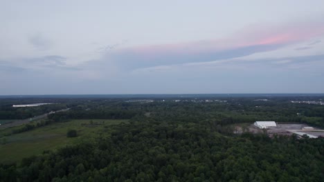 Aerial-drone-forward-moving-shot-over-the-lush-green-forest-beside-a-road-during-evening-time-after-sunset