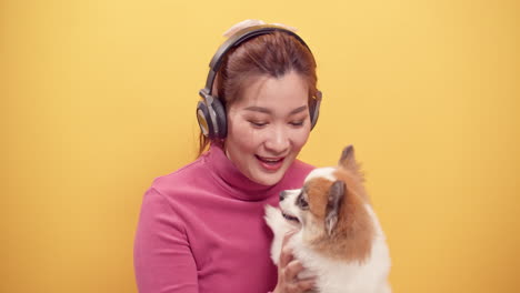 Wunderschöne-Asiatische-Frau-Verwendet-Ein-Digitales-Tablet-Und-Eine-Streaming-anwendung,-Um-Fröhlich-Musik-über-Kopfhörer-Zu-Hören,-Während-Sie-Den-Hund-Zur-Entspannung-Auf-Hellgelbem-Hintergrund-Spielt-1