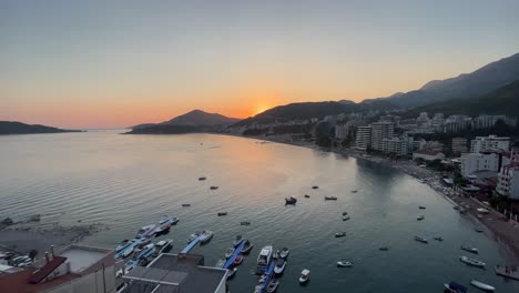 Lovely-sunset-over-rafailovici-in-montenegro,-high-flying-drone-shot-in-the-evening-on-a-sunny-day-during-sunset,-sun-setting-over-the-mountain-with-boats-and-cars-in-the-view