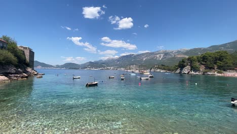 Blaues-Wasser-Und-Boote-Rund-Um-Sveti-Stefan-In-Montenegro-Mit-Hohen-Bergen-Im-Hintergrund,-Ein-Sehr-Berühmter-Strand,-An-Dem-Alle-Aktuellen-Prominenten-Hingehen,-Wenn-Sie-Einen-Schönen-Urlaub-Verbringen-Möchten