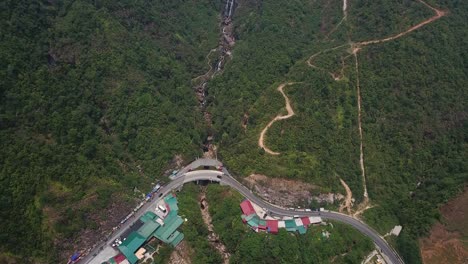A-distant-aerial-view-directly-above-a-winding-country-road-and-waterfall-in-Sapa,-Vietnam