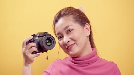 Happy-exuberant-Asian-woman-in-pink-clothing-using-a-digital-camera-against-an-isolated-yellow-background-1