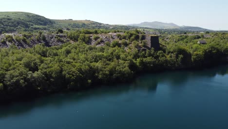 Aerial-view-Welsh-woodland-valley-slate-mining-shaft-and-quarry-lake-panning-across-Snowdonia-mountains