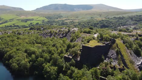 Aerial-view-Welsh-woodland-valley-slate-mining-shaft-and-Snowdonia-mountains-revealing-quarry-lake