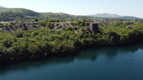Aerial-view-Welsh-woodland-valley-slate-mining-shaft-and-quarry-lake-under-Snowdonia-mountains