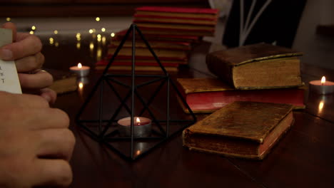 Several-candles-are-lit-on-the-table,-an-iron-pyramid-and-antique-books-are-visible