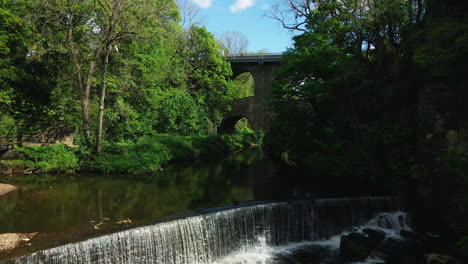 Peaceful-waterfall-in-British-river-with-green-trees-and-lush-grass-under-archway-aerial-slow-track-in-Peak-District