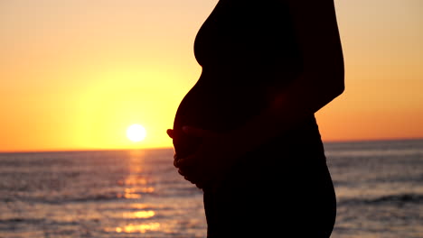 Close-up-Silhouette-of-pregnant-woman-touching-belly-in-front-of-tranquil-ocean-during-golden-sunset-light