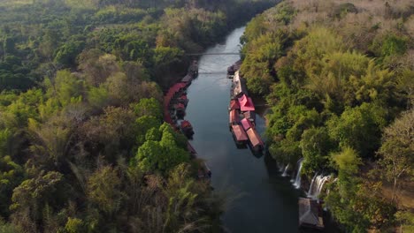 A-rotational-drone-shot-of-a-river-in-Thailand-in-the-middle-of-the-jungle,-with-waterfalls-and-a-floating-village-on-the-side-of-the-river